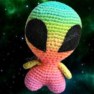 May include: A rainbow crochet alien with black eyes. The alien is standing against a green and black starry background.