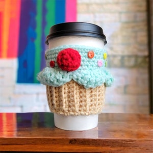 Pode incluir: Um protetor de café de cupcake de crochê em azul claro e bege com um pompom vermelho no topo e botões coloridos.