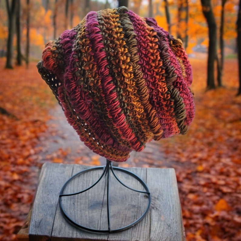 May include: A crocheted beanie hat in shades of brown, red, and orange. The hat is displayed on a black metal stand against a backdrop of autumn leaves.