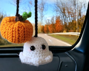 Op de afbeelding: Een gehaakte pompoen en een spook die aan een achteruitkijkspiegel in een auto hangen. De pompoen is oranje en het spook is wit met zwarte ogen.