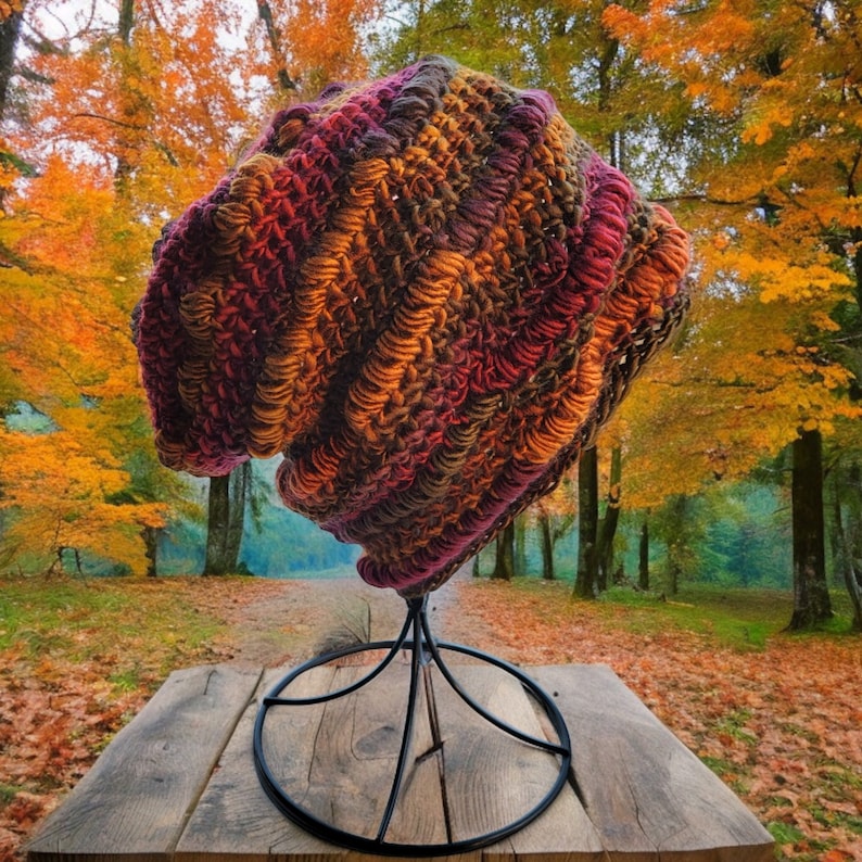May include: A crocheted beanie hat in shades of burgundy, brown, and orange. The hat is displayed on a black metal stand against a backdrop of fall foliage.