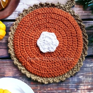 May include: A crocheted pumpkin pie hot pad with a brown crust and a white center. The hot pad is on a wooden surface.