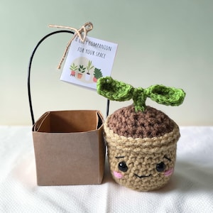 Pode incluir: Uma planta de crochê com um rosto sorridente e folhas verdes, ao lado de uma pequena caixa marrom e um cartão que diz "A Cozy Companion For Your Space". A planta está em um vaso marrom com uma borda marrom clara.
