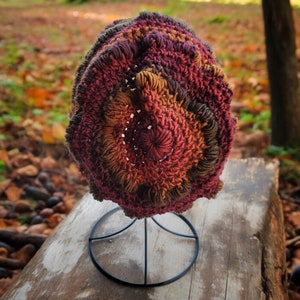 May include: A crocheted hat with a multi-colored design of brown, red, and orange. The hat is displayed on a black metal stand.