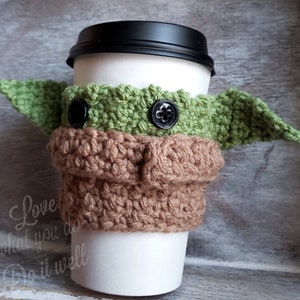 May include: A crocheted coffee cup cozy in the shape of a green alien character with large ears. The cozy has brown and green yarn and black buttons for eyes. The text "Love what you do Do it well" is printed on the cup.