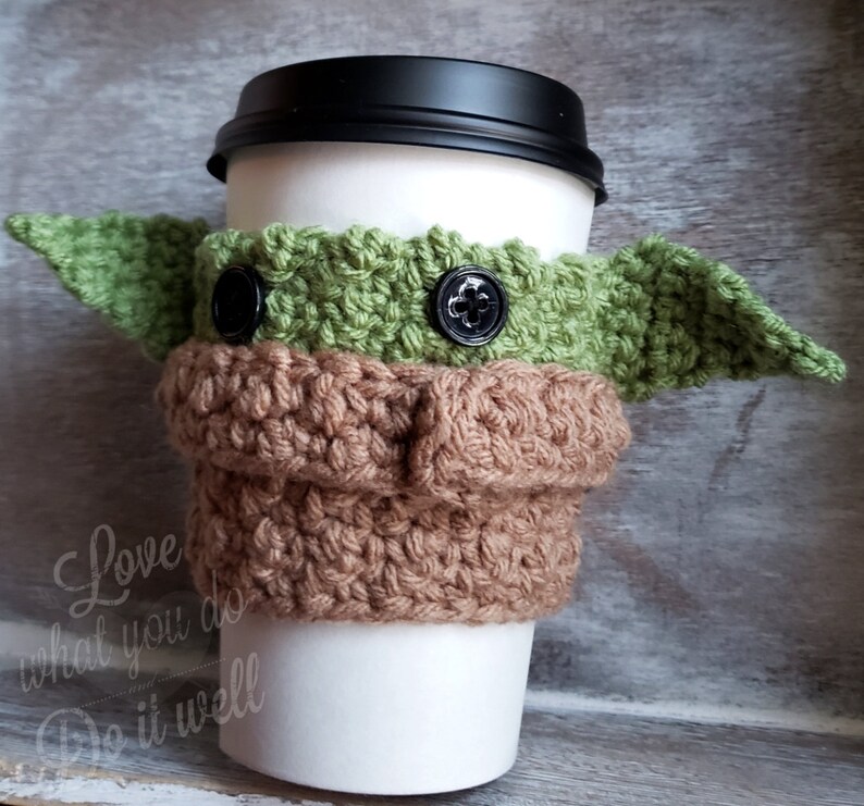May include: A crocheted coffee cup cozy in the shape of a green alien character with large ears. The cozy has brown and green yarn and black buttons for eyes. The text "Love what you do Do it well" is printed on the cup.