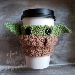 May include: A crocheted coffee cup cozy in the shape of a green alien character with brown ears and black buttons for eyes. The cozy is on a white to-go cup with a black lid. The text "Love what you do Do it well" is printed on the cup.