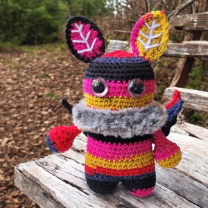 May include: A handmade crochet creature with large, clear eyes and a fluffy gray collar. The body is striped with vibrant colors: pink, yellow, red, and black. The ears are leaf-shaped with white detailing. The creature is sitting on a wooden surface.