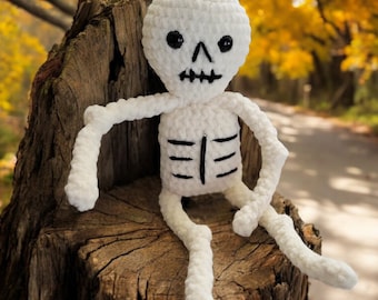 Puede incluir: Un muñeco de esqueleto de crochet blanco sentado en un tocón de árbol. El muñeco tiene ojos negros y una boca negra. El muñeco lleva un atuendo de crochet blanco con detalles de costillas negras.