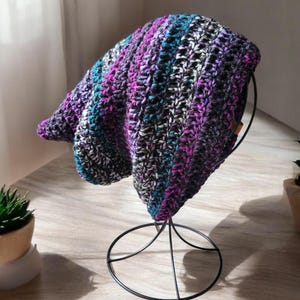 May include: A slouchy beanie hat crafted with a textured crochet stitch pattern. The yarn features a variegated color palette of deep purple, vibrant magenta, bright teal, and charcoal gray, creating a multi-colored striped effect throughout.