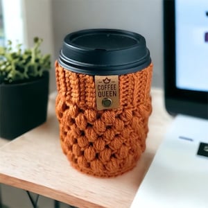 May include: A crocheted coffee cup cozy in a burnt orange color with a brown leather tag that says "Coffee Queen" with a crown above the word "Coffee".