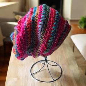May include: A colorful, handmade crochet hat displayed on a black metal stand. The hat features a vibrant mix of pink, blue, green, and orange yarns, creating a textured, striped pattern. The hat is a beanie style, perfect for cold weather.