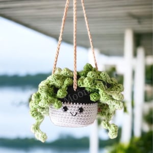 Pode incluir: Um vaso de plantas pendurado de crochê com um rosto sorridente e folhas verdes. O vaso é feito de fio branco e tem uma borda preta. As folhas são feitas de fio verde e são enroladas e torcidas.