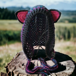 May include: A black crocheted cat hat with pink ears and long, colorful yarn tails. The hat is sitting on a tree stump.