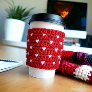 May include: A white disposable coffee cup with a black lid, featuring a red crocheted sleeve decorated with a repeating pattern of small white hearts. The sleeve is textured and fits snugly around the center of the cup, providing a decorative cover.