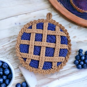May include: A crocheted pot holder shaped like a blueberry pie with a brown crust and a blue filling. The pot holder has a loop for hanging.