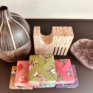 Puede incluir: Una colección de objetos decorativos: un jarrón a rayas blancas y negras, un bloque de piedra texturizada, una geoda púrpura y cajas de regalo de origami con diseños florales. Los objetos están dispuestos sobre una superficie oscura.