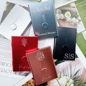 May include: A collection of personalized metal cardholders in silver, blue, red, and black. Each cardholder features a unique design, including floral patterns, names, and initials. The blue cardholder has the text "Chacha" and "Investment Manager".