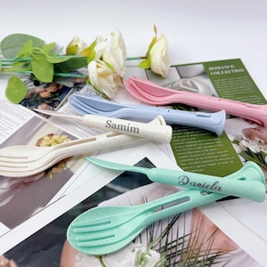 May include: A collection of colorful, personalized cutlery sets. The sets include a fork, spoon, and knife in pastel shades of pink, blue, and green, with names engraved on some pieces. White flowers and printed paper are in the background.