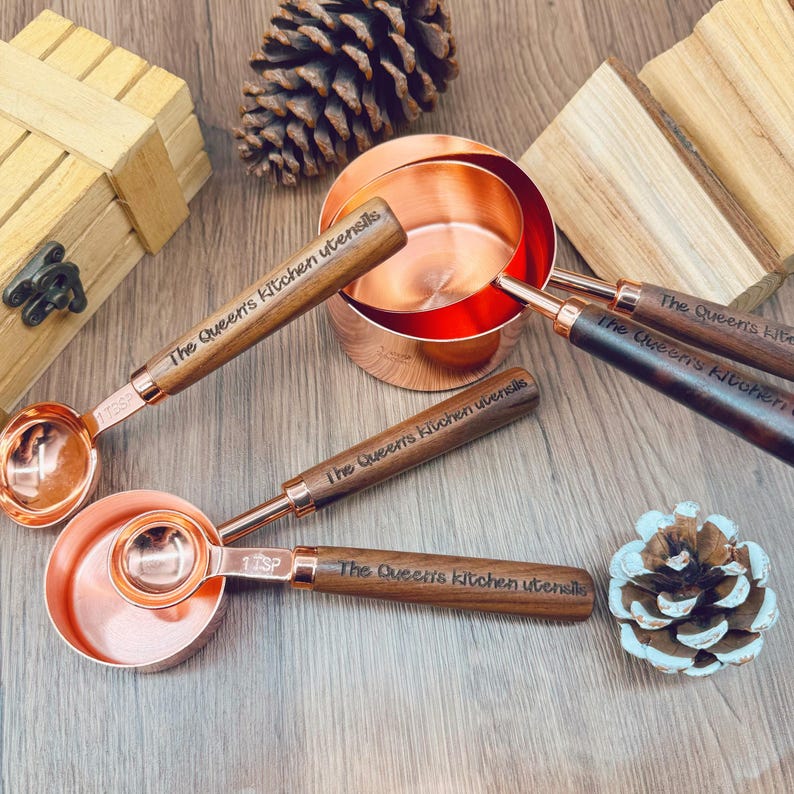 May include: A set of copper measuring cups and spoons with wooden handles. The text "The Queen's Kitchen Utensils" is engraved on the handles. The set includes a measuring cup and several measuring spoons, with "1 TSP" on one spoon.