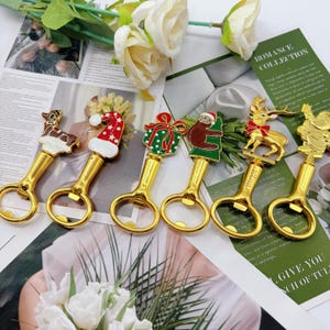 May include: A collection of gold-colored bottle openers with festive holiday-themed designs. Each opener features a different charm, including a reindeer, Santa hat, gift, sloth, reindeer, and Santa Claus. The openers are arranged on a printed background.