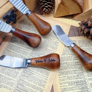 May include: Four cheese spreaders with stainless steel blades and wooden handles are arranged on a vintage book page. One spreader has the name "Velas Catoru" engraved on the handle. A pinecone and wooden block are in the background.