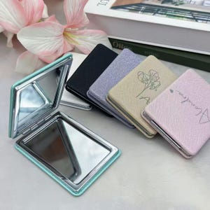 May include: A collection of rectangular compact mirrors with textured covers in black, lavender, beige, and light pink. One beige mirror features a floral line drawing, while the pink one displays the name Alondra. An open teal mirror shows two reflective surfaces.