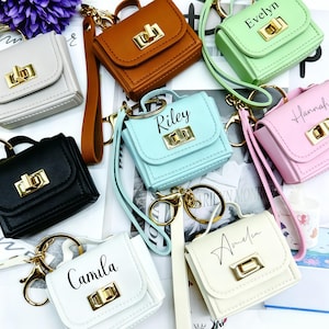 Puede incluir: Una colección de llaveros de bolsos en miniatura en varios colores, incluyendo negro, blanco, marrón, verde claro, azul claro y rosa. Cada bolso cuenta con un cierre giratorio dorado, un asa superior y una correa de muñeca. Algunos bolsos están personalizados con nombres como Evelyn, Riley, Hannah, Camila y Amelia en letra negra.