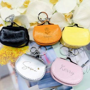 May include: A selection of coin purses in black, orange, yellow, silver, and pink. Each purse has a silver keychain and is personalised with a name. The orange purse has a rose design and the name "Victoria".