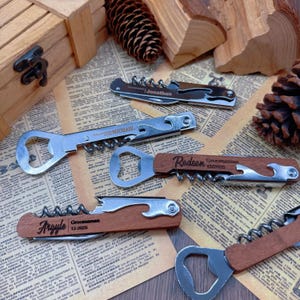May include: Several personalized corkscrew and bottle opener tools are displayed. Some tools have wooden handles with engraved names and dates. The tools are silver and brown, with a corkscrew, bottle opener, and knife. The background includes a wooden box, pine cones, and newspaper.