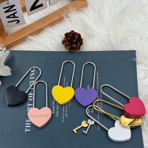 Peut inclure: Plusieurs cadenas en forme de cœur de couleurs variées : noir, rose, jaune, violet, doré et blanc. Chaque cadenas est doté d'une anse argentée. Certains cadenas sont gravés de noms. Deux petites clés sont également présentes.