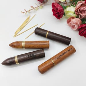 Peut inclure: Trois porte-cure-dents en bois avec des accents dorés. Les porte-cure-dents sont en forme de balles et ont des textes gravés différents : "Billy", "60/70 birthday cruise" et un porte-cure-dents vide.