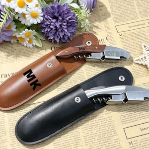 May include: Two stainless steel wine openers with corkscrews, one featuring a wood-grain handle with the letters MK, and one with a solid silver-tone handle. Each opener includes a matching leather-style storage sleeve, one tan and one black.
