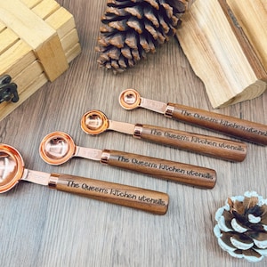 May include: A set of four copper-colored measuring spoons with wooden handles. Each spoon is engraved with "The Queen's Kitchen utensils". The spoons are arranged on a wooden surface, with a pine cone and wooden box in the background.