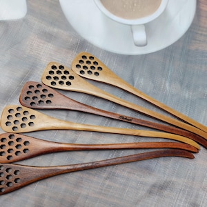 May include: Six wooden honey dippers with honeycomb designs. The dippers are of varying lengths and have a natural wood finish. The dippers are arranged in a fan shape with the longest dipper at the top. The dipper at the bottom has the word "Michael" engraved on it.