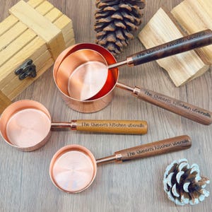 May include: Copper measuring cups with wooden handles. The handles are engraved with "The Queen's Kitchen utensils". The cups are in various sizes and are arranged on a wooden surface with a pine cone and wood pieces.