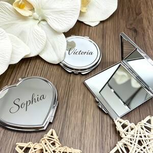 Pode incluir: Três espelhos de bolso em metal prateado. Um tem formato de coração com o nome Sophia gravado, um é redondo com o nome Victoria gravado e um é retangular e está aberto, revelando dois espelhos internos. As superfícies são reflexivas e polidas.
