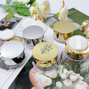 May include: An assortment of cosmetic containers with metallic silver and gold lids. One container has a floral design and the name "Emma" printed on the lid. The containers are clear with a white powder puff in one.