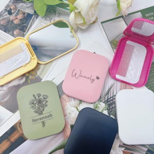 Può includere: Una collezione di specchietti compatti e set di pettini in vari colori, tra cui giallo, rosa, verde, blu navy e bianco. Alcuni compatti presentano testi come "Waverly" e "Savannah". Gli specchietti sono aperti, rivelando la superficie riflettente e un pettine.