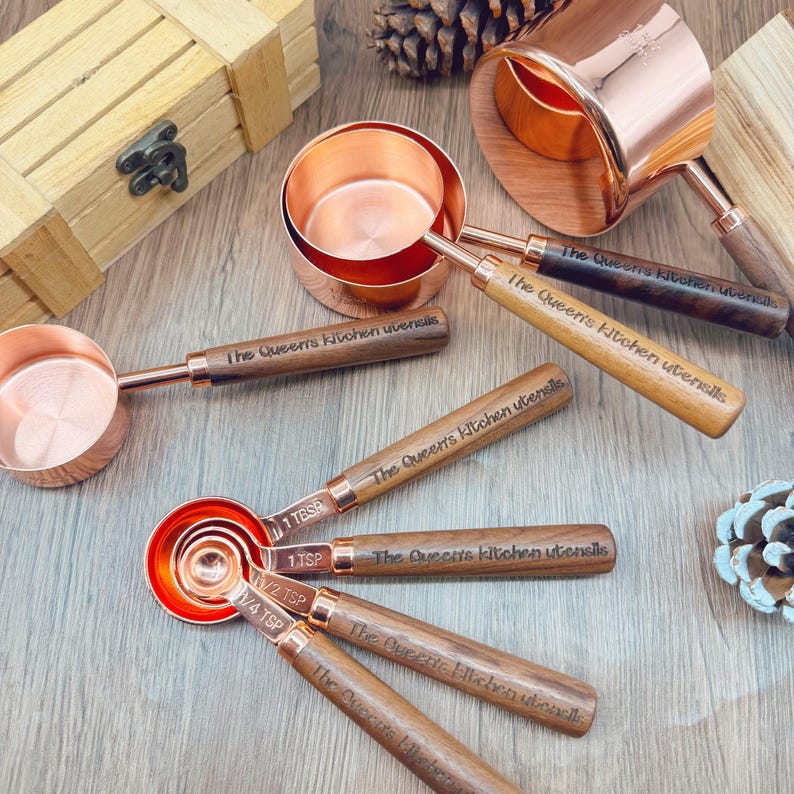 May include: Copper measuring cups and spoons with wooden handles. The text "The Queen's Kitchen Utensils" is engraved on the handles. The set includes various sizes, from 1/4 tsp to 1 cup. The copper has a shiny finish.