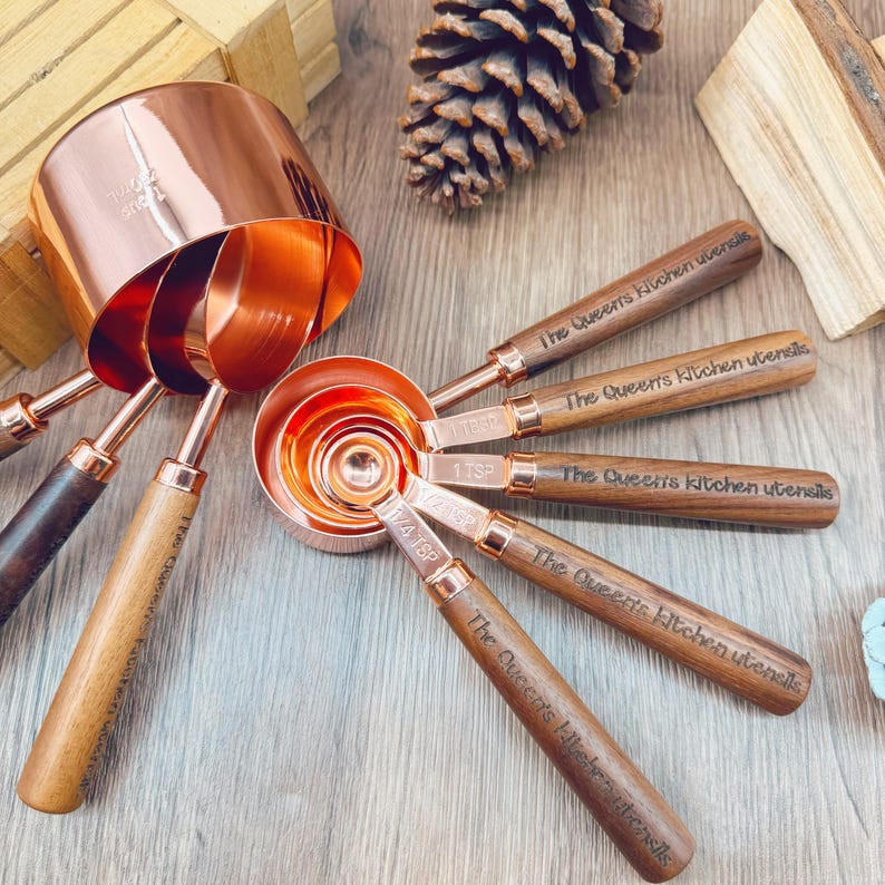 May include: A set of copper measuring cups and spoons with wooden handles. The spoons are engraved with "The Queen's Kitchen Utensils". The set includes various sizes for accurate measurements in cooking and baking.