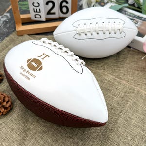 May include: Two white footballs with brown and white stitching. One football has the initials "JT", a football graphic, and the text "Ring Bearer 5/30/2026" printed on it. The footballs are on a textured, tan surface.