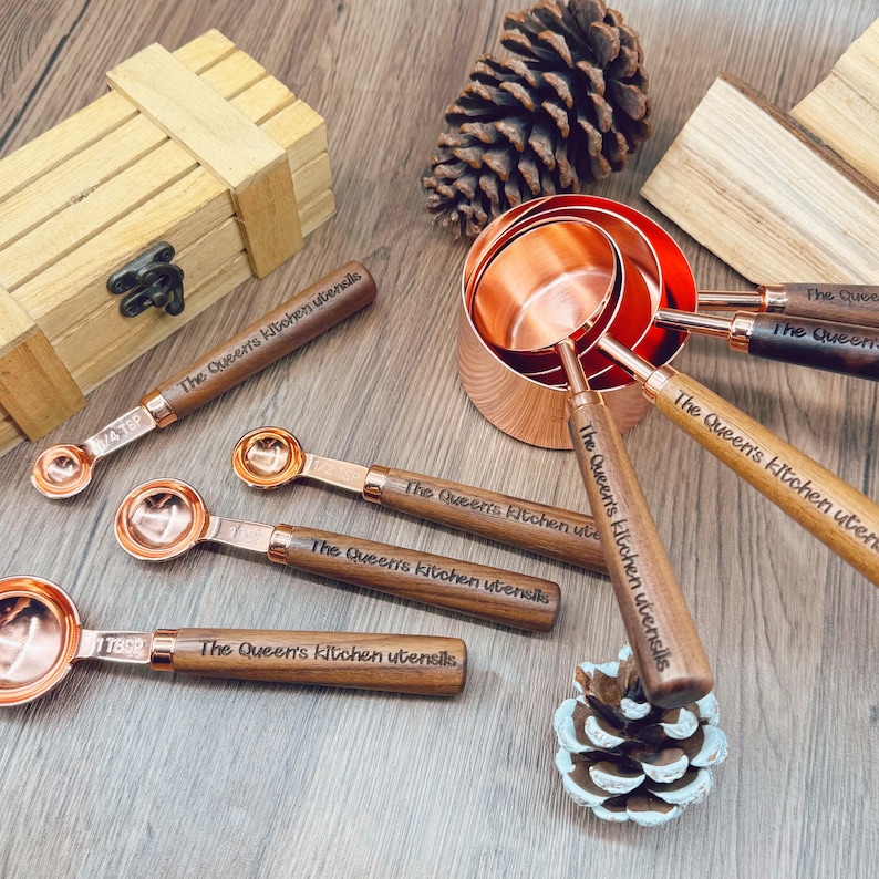 May include: A set of copper measuring cups and spoons with wooden handles. The spoons and cups are arranged on a wooden surface, with a wooden box and pinecone in the background. The text "The Queen's Kitchen Utensils" is engraved on the handles.