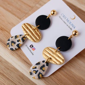 Peut inclure: Paire de boucles d'oreilles faites à la main avec un design unique. Chaque boucle d'oreille comprend un cercle noir, un ovale doré texturé et une forme de goutte avec un motif léopard. Les boucles sont présentées sur une carte blanche.