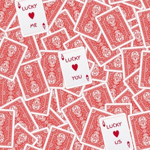 Op de afbeelding: Een verzameling rode en witte speelkaarten met een schedelpatroon op de achterkant. Drie kaarten liggen open en tonen de tekst LUCKY ME, LUCKY YOU en LUCKY US, elk met een aassymbool en een rood hart in het midden van de kaart.