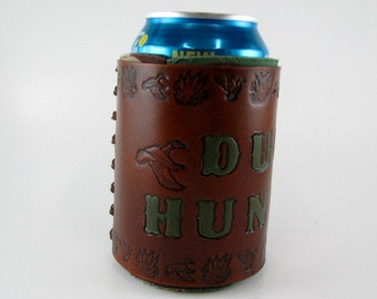May include: Brown leather can cooler with the words "DUCK HUNT" in green lettering. The cooler features a duck graphic and decorative embossed duck and plant designs. A blue can is inside the cooler.