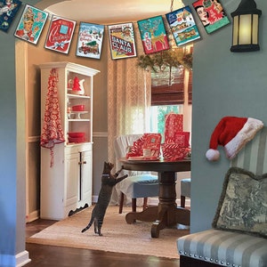 Pu&ograve; includere: Una ghirlanda natalizia con carte in stile vintage che mostrano immagini di Babbo Natale, renne e un albero di Natale. La ghirlanda &egrave; appesa in una stanza con un gatto, un tavolo e una sedia.