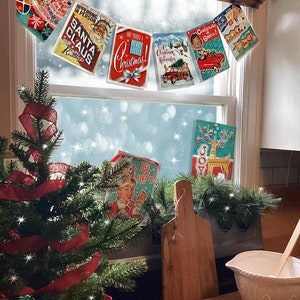 Pu&ograve; includere: Una collezione di cartoline di Natale vintage appese a una finestra con un piccolo albero di Natale e utensili da forno su un bancone di legno sottostante. Le cartoline presentano immagini e testi festivi come "Let Your Christmas Spirit Shine!" e "Merry Christmas!"