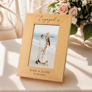 Peut inclure: Un cadre photo en chêne clair avec le mot "Engaged" en écriture cursive et un symbole de cœur. Le cadre contient une photo d'un couple dansant sur une plage. Les noms "KLEIN & ELAINE" et la date "26.08.2025" sont imprimés en bas.