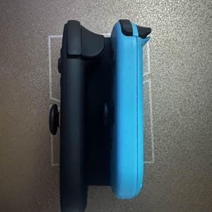 Puede incluir: Un mando de juego negro y azul claro están unidos a un soporte negro. Los mandos tienen botones y joysticks negros. El fondo es una superficie gris texturizada.